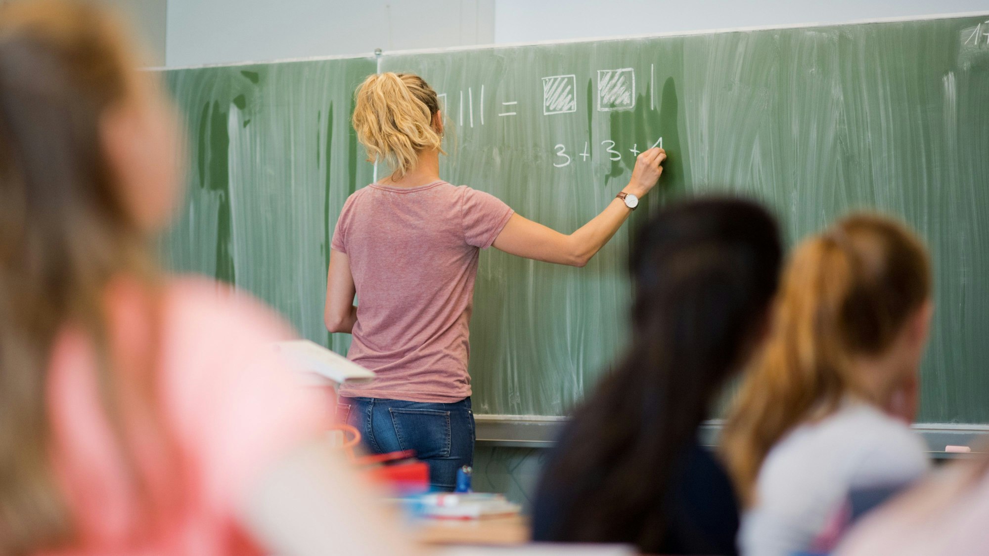 Eine Lehrerin schreibt vor Schülerinnen eine Mathe-Aufgabe an die Tafel.