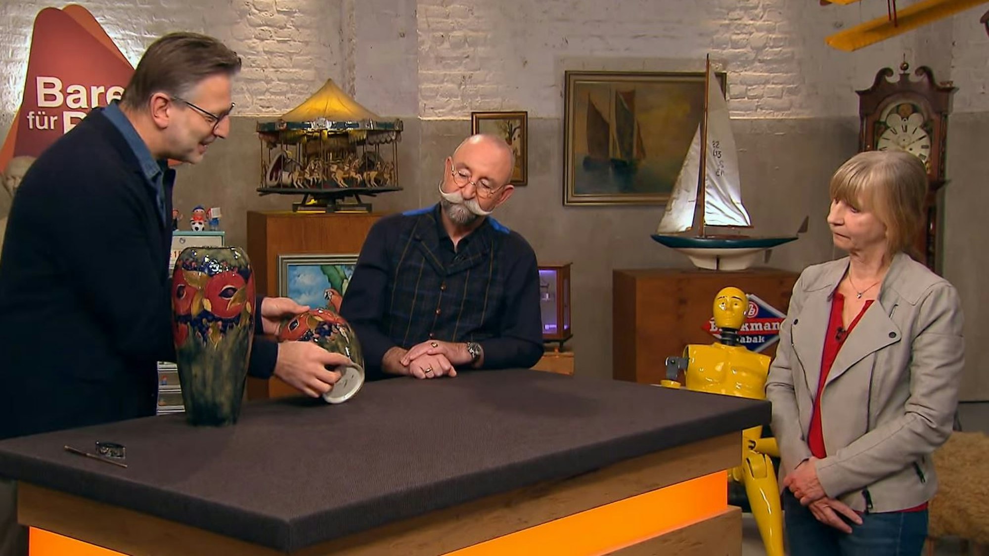 Experte Detlev Kümmel (l.) bei der Expertise bei „Bares für Rares“, Moderator Horst Lichter (M.) und Verkäuferin Petra Busch-Gremmler (r.) lauschen gespannt.