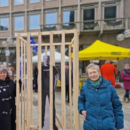 Zwei Frauen stehen zwischen einem eingegitterten Plakat von Julian Assange.