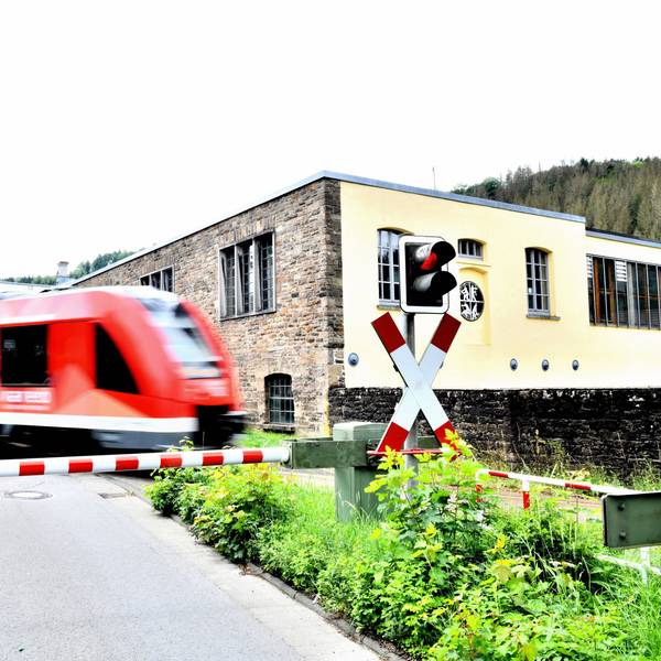 Ein Zug der Regionalbahn 25 passiert den Bahnübergang Märkische Straße in Engelskirchen, im Hintergrund die Fabrik Ermen und Engels, wo heute das Industriemuseum und das Rathaus zu finden sind.