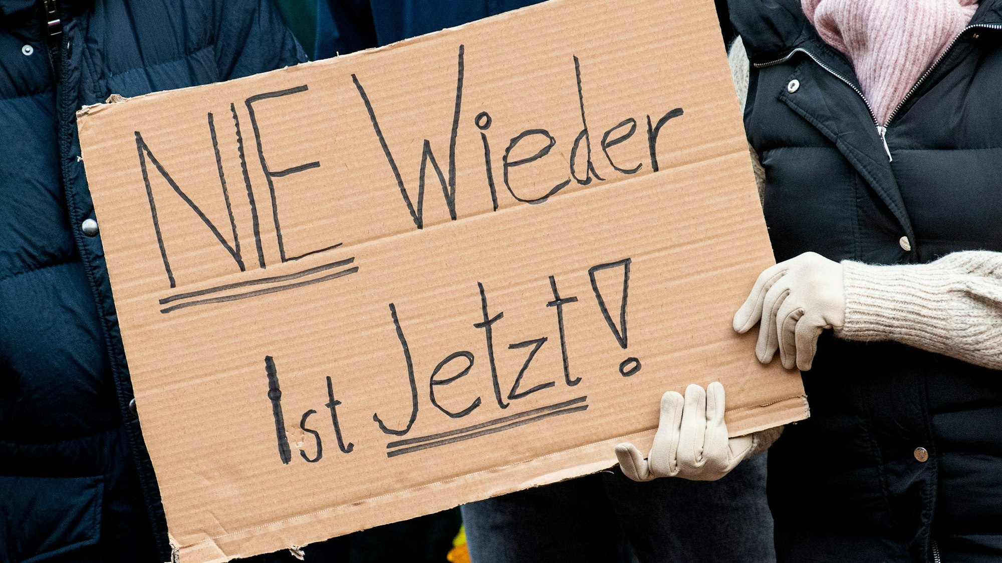 Teilnehmer einer Demonstration gegen Rechtsextremismus halten ein Schild „Nie wieder ist jetzt" in Händen.