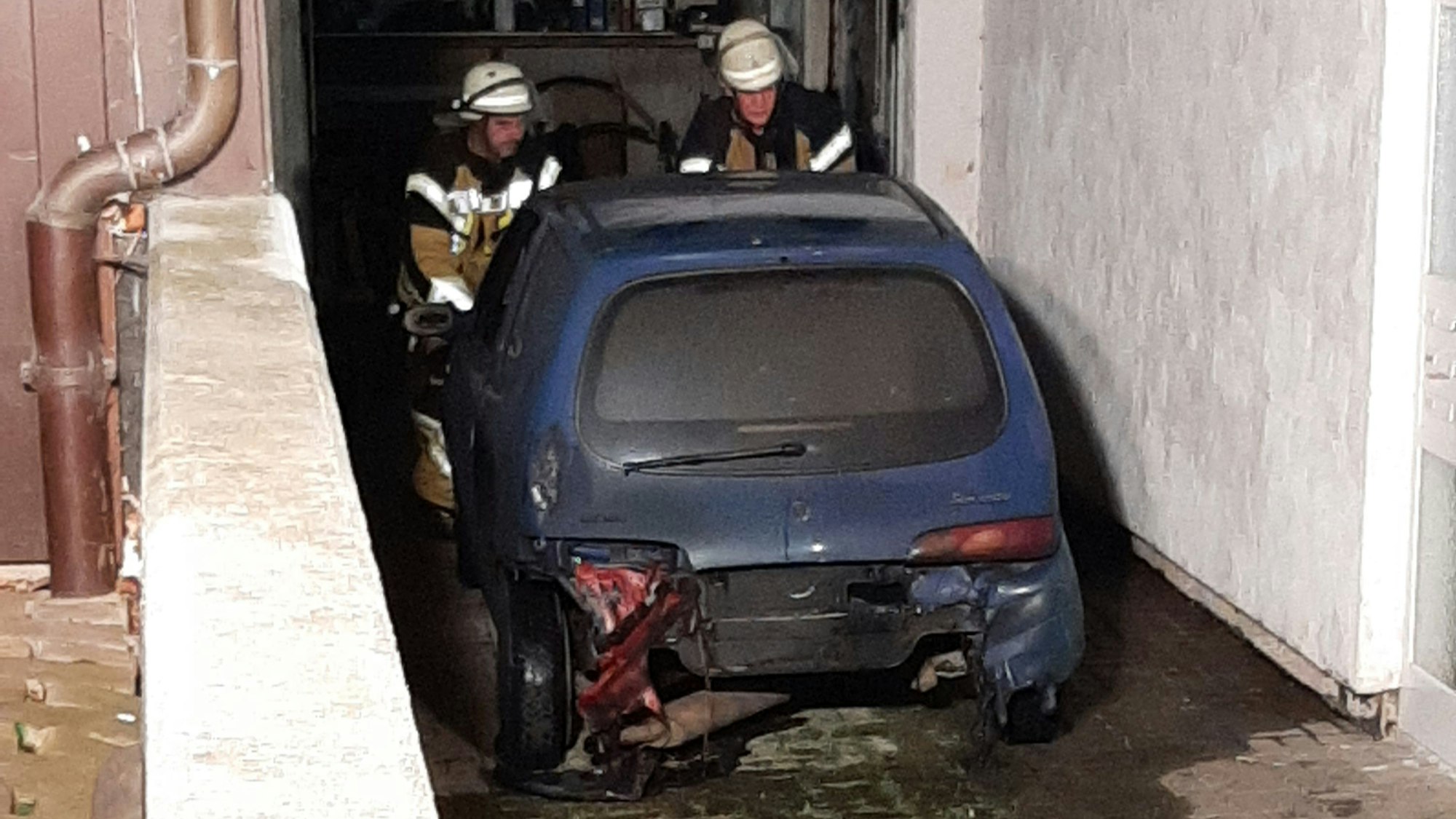 Nach einem Brand in einer Garage in Kall stehen zwei Feuerwehrleute an einem blauen Kleinwagen.