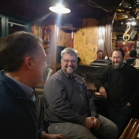 Drei Männer sitzen in einem Pub, vor ihnen stehen Biergläser auf dem Tisch, sie lachen.