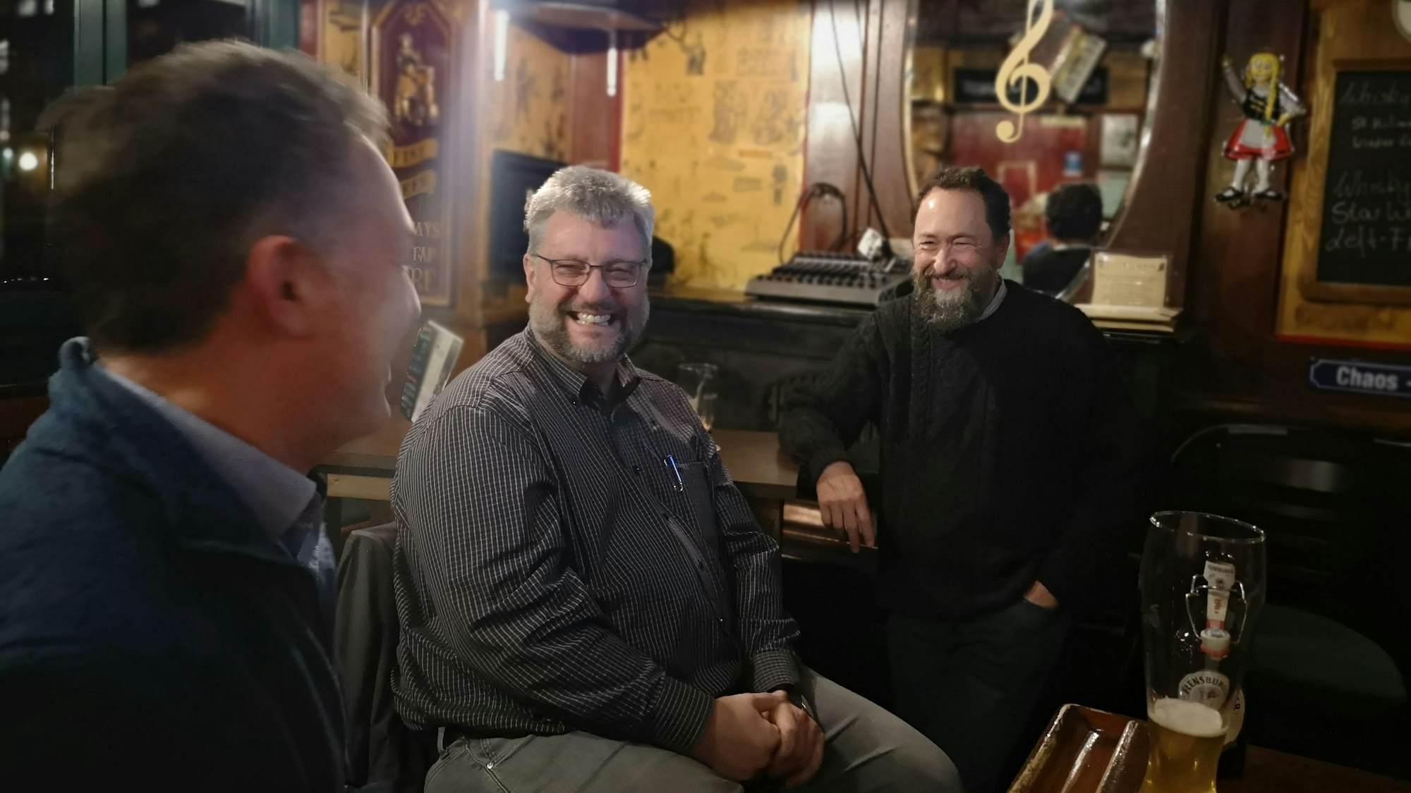 Drei Männer sitzen in einem Pub, vor ihnen stehen Biergläser auf dem Tisch, sie lachen.