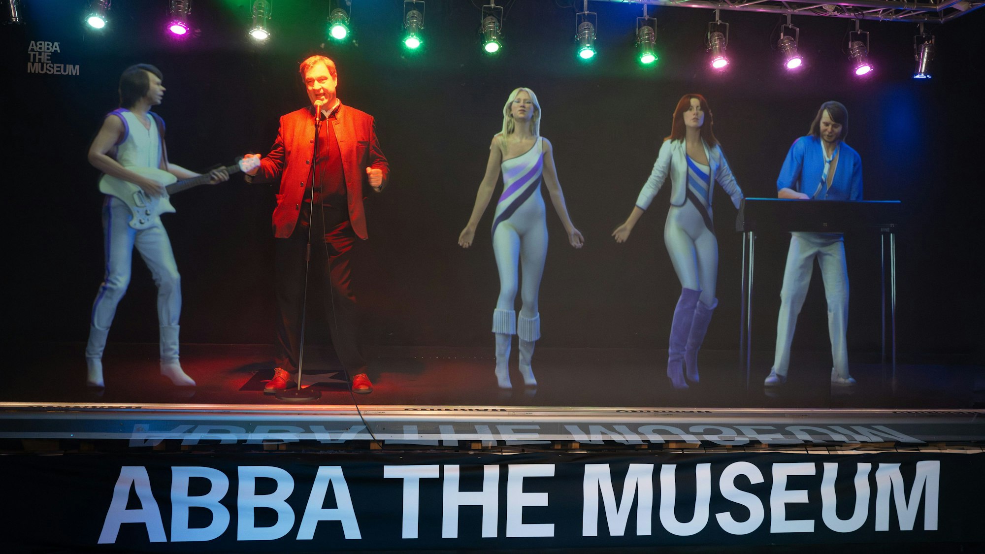 Bayerns Ministerpräsident Söder steht auf einer Bühne. Er singt und tanzt mit ABBA-Hologrammen.
