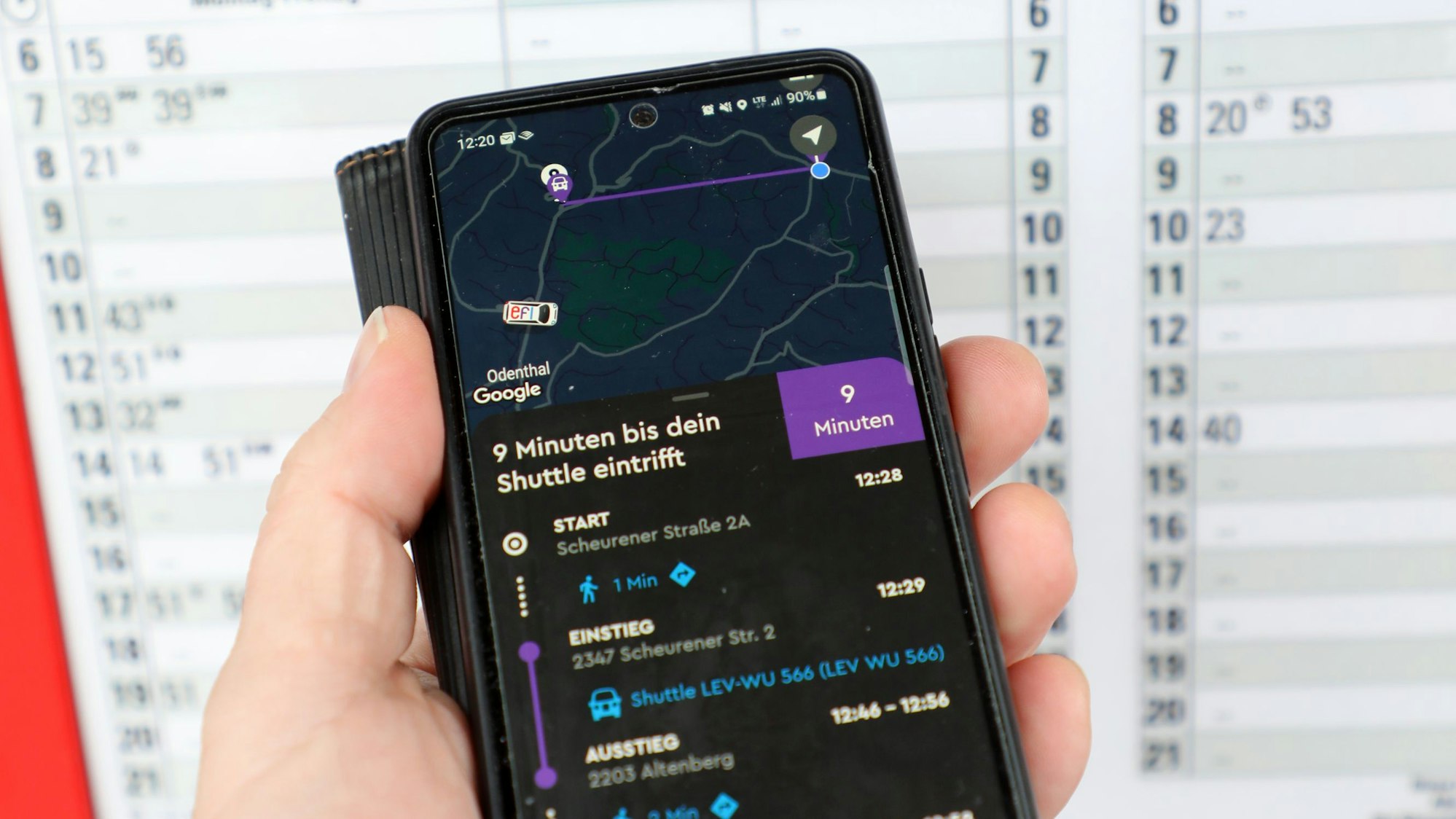 Eine Hand hält ein Handy vor einem Busfahrplan.