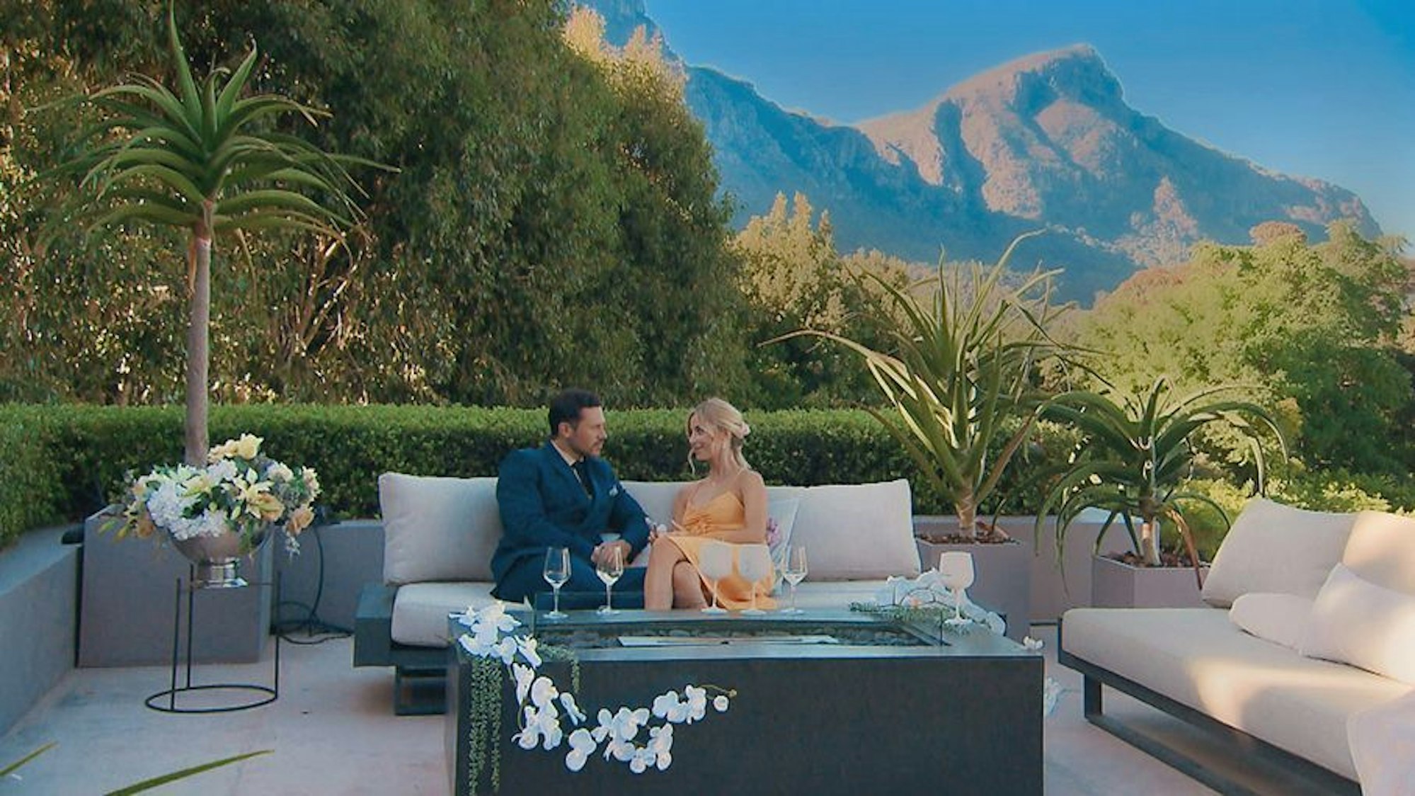 Ein Mann und eine Frau sitzen auf einer Couch. Hinter ihnen sind Berge zu sehen.