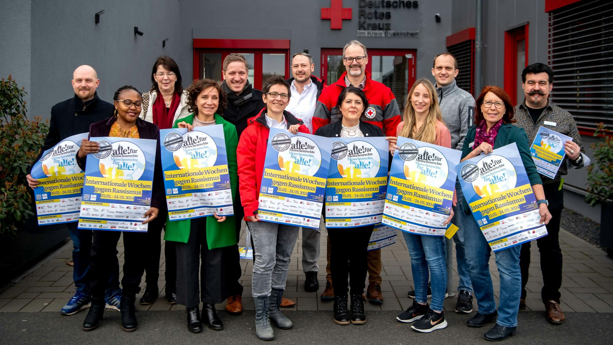 Eine Gruppe Männer und Frauen steht vor dem DRK-Zentrum in Euskirchen. Einige halten Plakate in den Händen, die auf die Aktionswochen gegen Rassismus hinweisen.