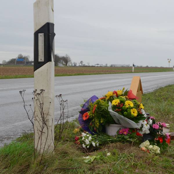 Das Bild zeigt die L163. Im Hintergrund fahren Autos, im Vordergrund steht ein Holzkreuz, das fast vollständig von Blumen verdeckt ist.