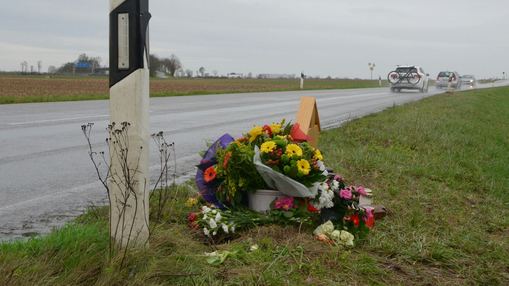 Das Bild zeigt die L163. Im Hintergrund fahren Autos, im Vordergrund steht ein Holzkreuz, das fast vollständig von Blumen verdeckt ist.