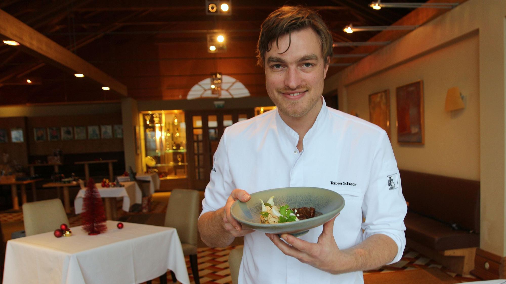 Torben Schuster zeigt seine köstlichen Kreationen liebevoll auf dem Teller auf. Er lächelt in Richtung der Kamera, befindet sich in dem von ihm geleiteten Restaurant Gut Lärchenhof in Kerpen.