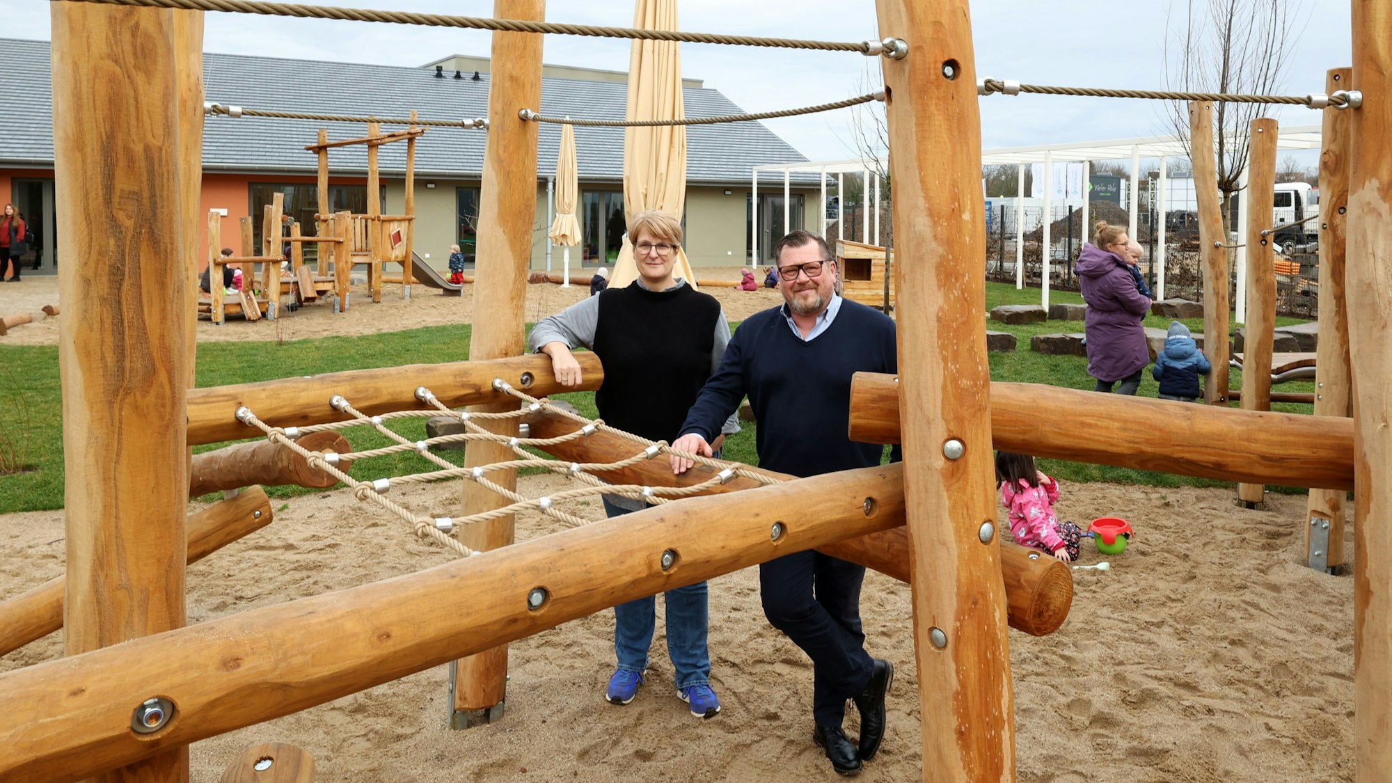 Zu sehen sind eine Frau und ein Mann auf einem Außengelände einer Kindertagesstätte, die durch ein Klettergerüst hindurch in die Kamera blicken.