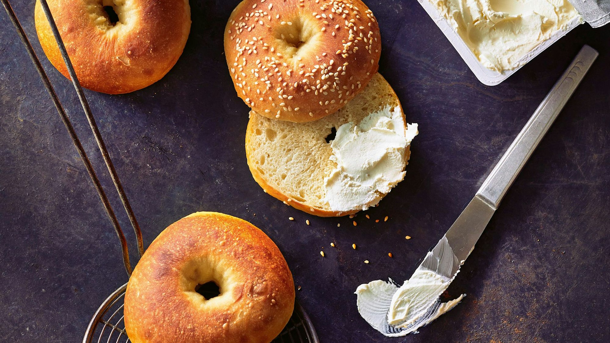 Bagels liegen auf einer Ablage, einer ist aufgeschnitten und zum Teil mit Frischkäse beschmiert.