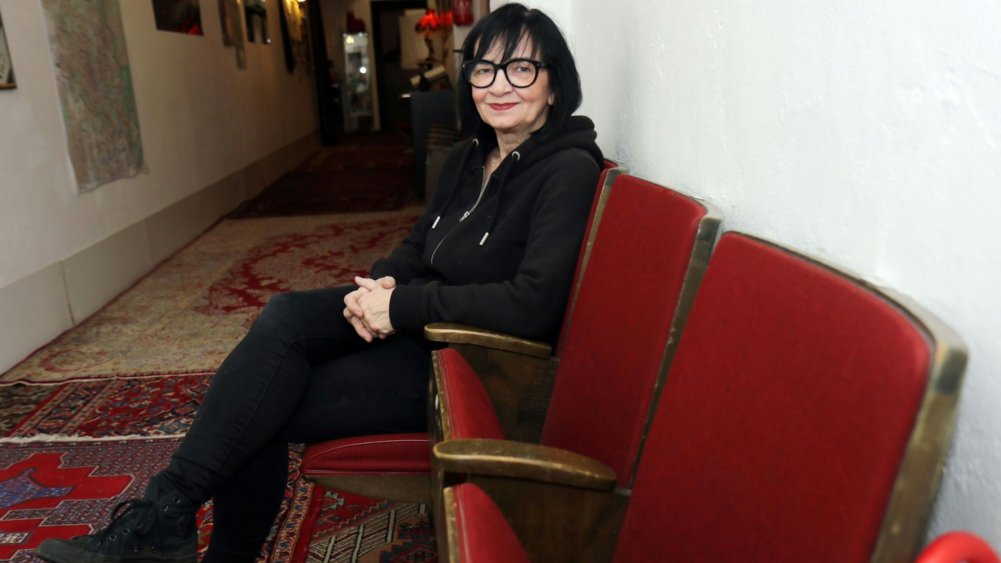 Frau mit schwarzen Haaren, schwarzer Brille und schwarzer Kleidung sitzt auf einem roten Sesel in einem Flur und schaut in die Kamera