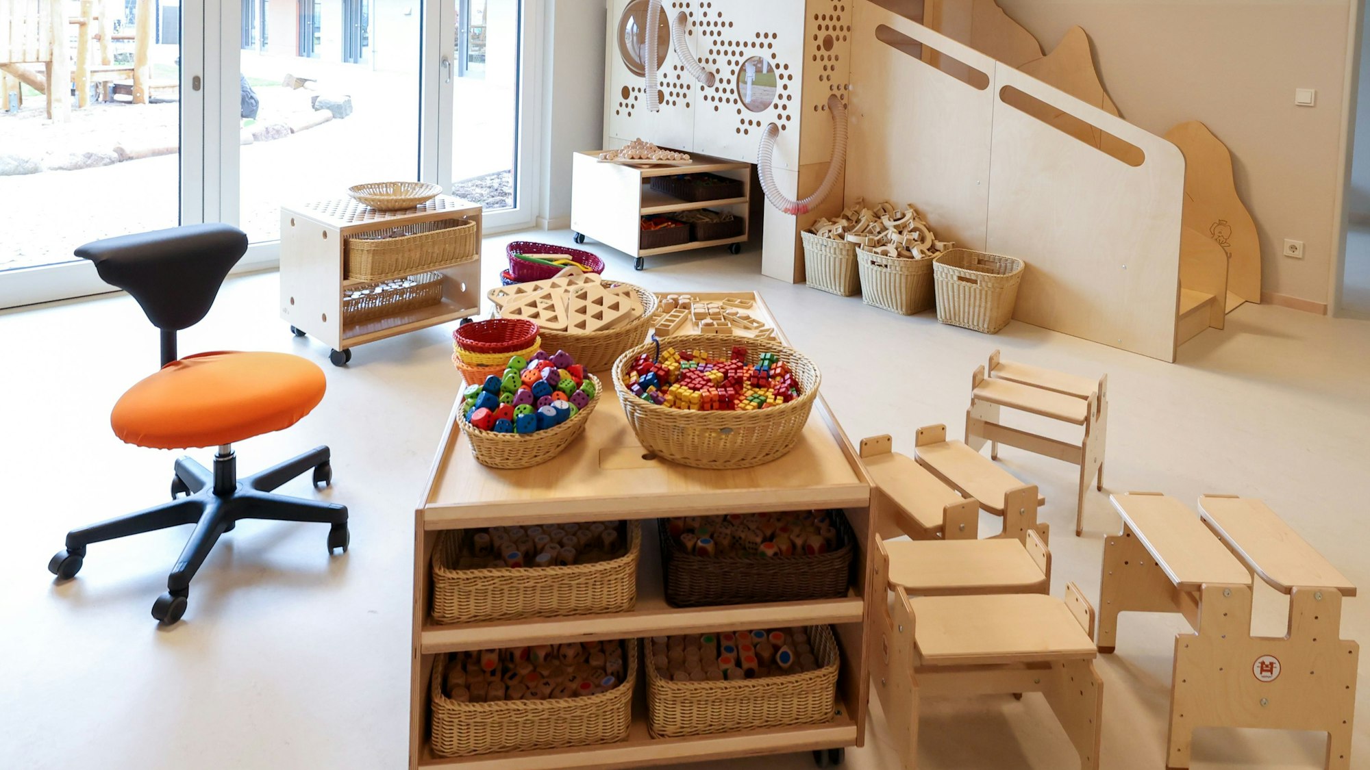 Zu sehen ist ein Gruppenraum in einer Kita mit Holz-Spielzeug, Tischen und Stühlen.