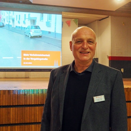 Ein Mann steht vor einer Bühne, an der Wand ist eine Präsentation zu sehen.