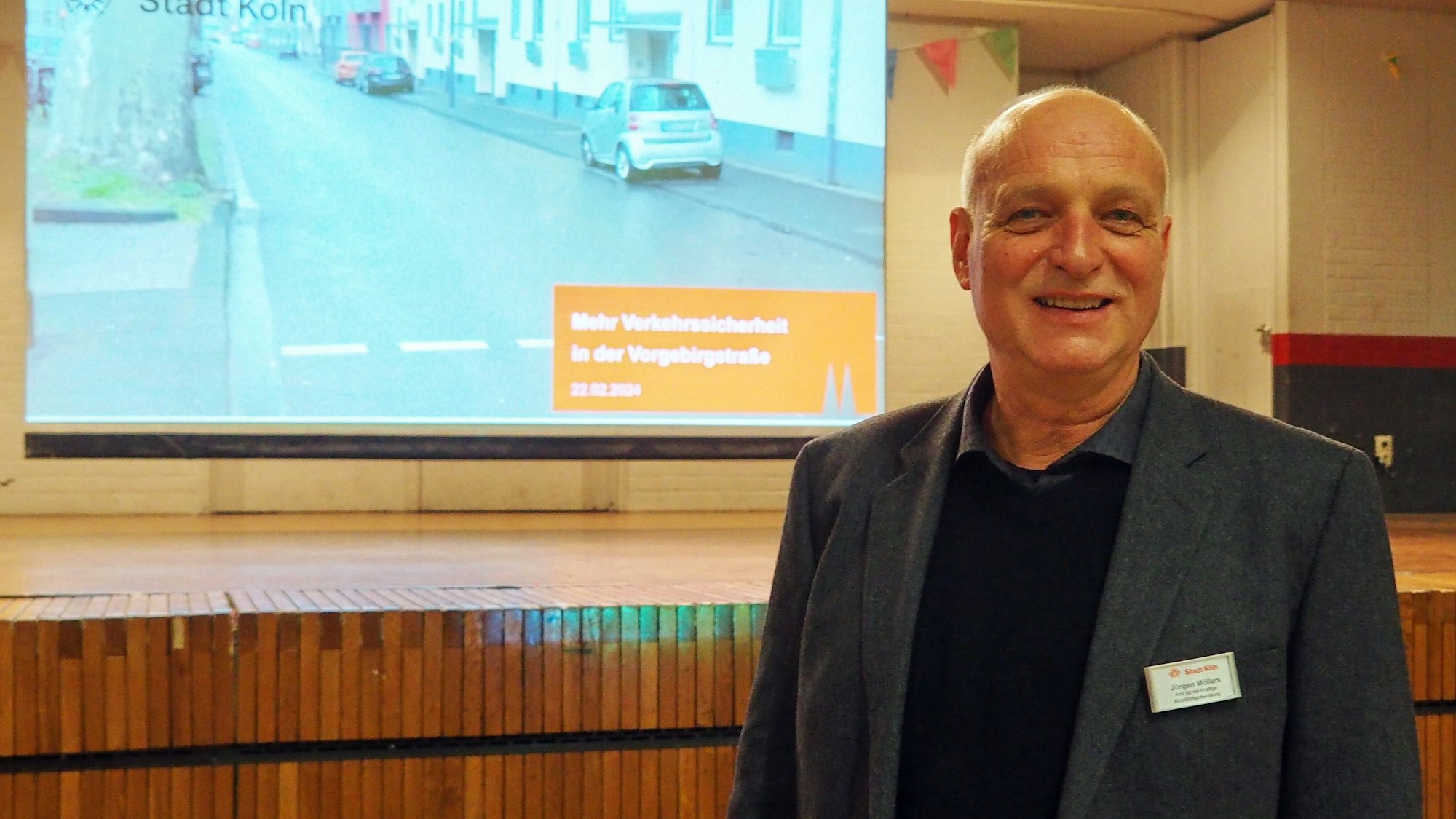 Ein Mann steht vor einer Bühne, an der Wand ist eine Präsentation zu sehen.