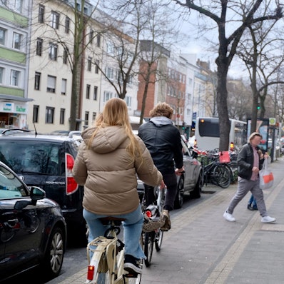 Radfahrer fahren auf dem Radstreifen, Fußgänger sind auf dem direkt daneben liegenden Gehweg unterwegs.