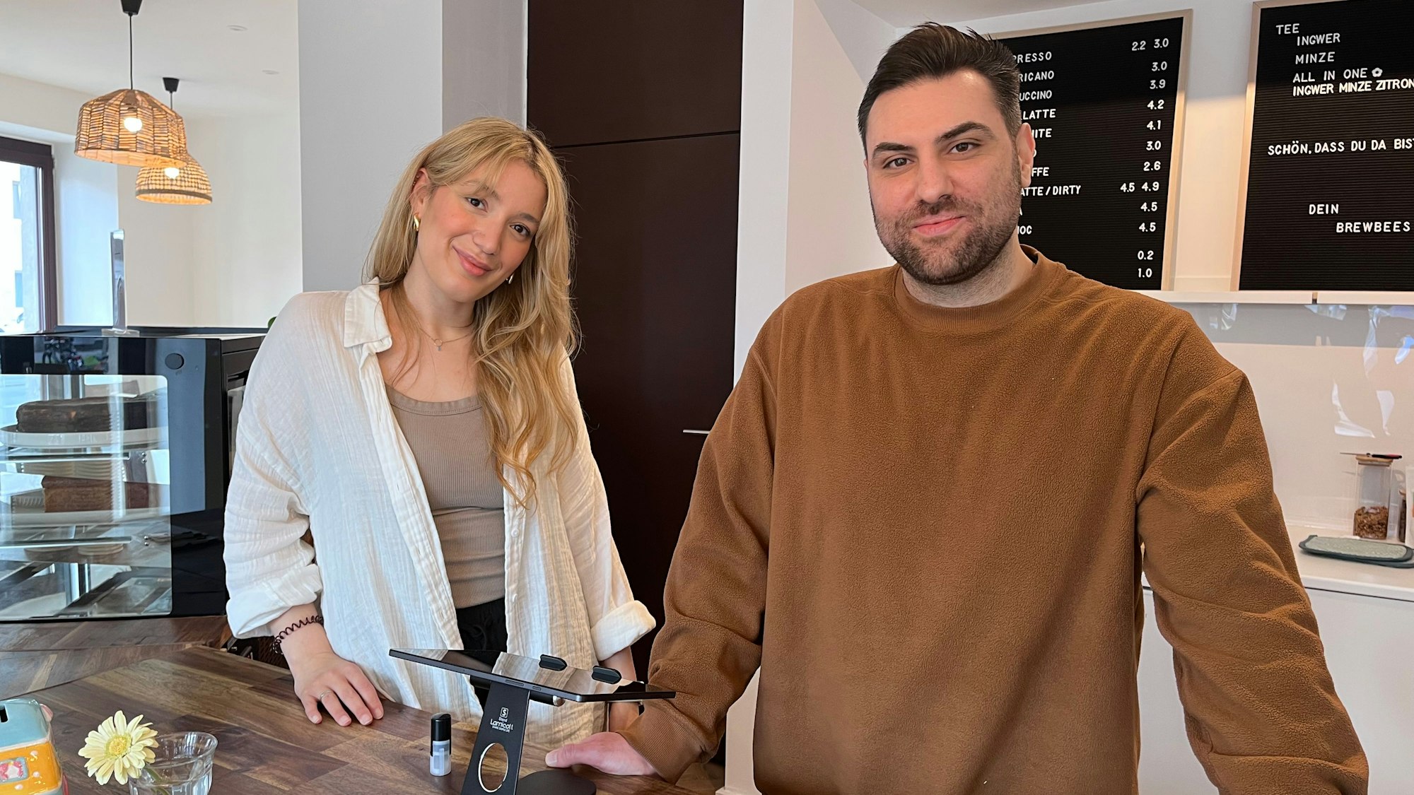 Eine Frau mit blondem Haar und ein Mann mit kurzen dunklen Haaren stehen hinter der Theke in einem Café.