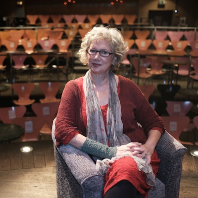 Eine Frau mittleren Alters sitzt auf der Bühne in einem kleinen Theater vor Stuhlreihen.