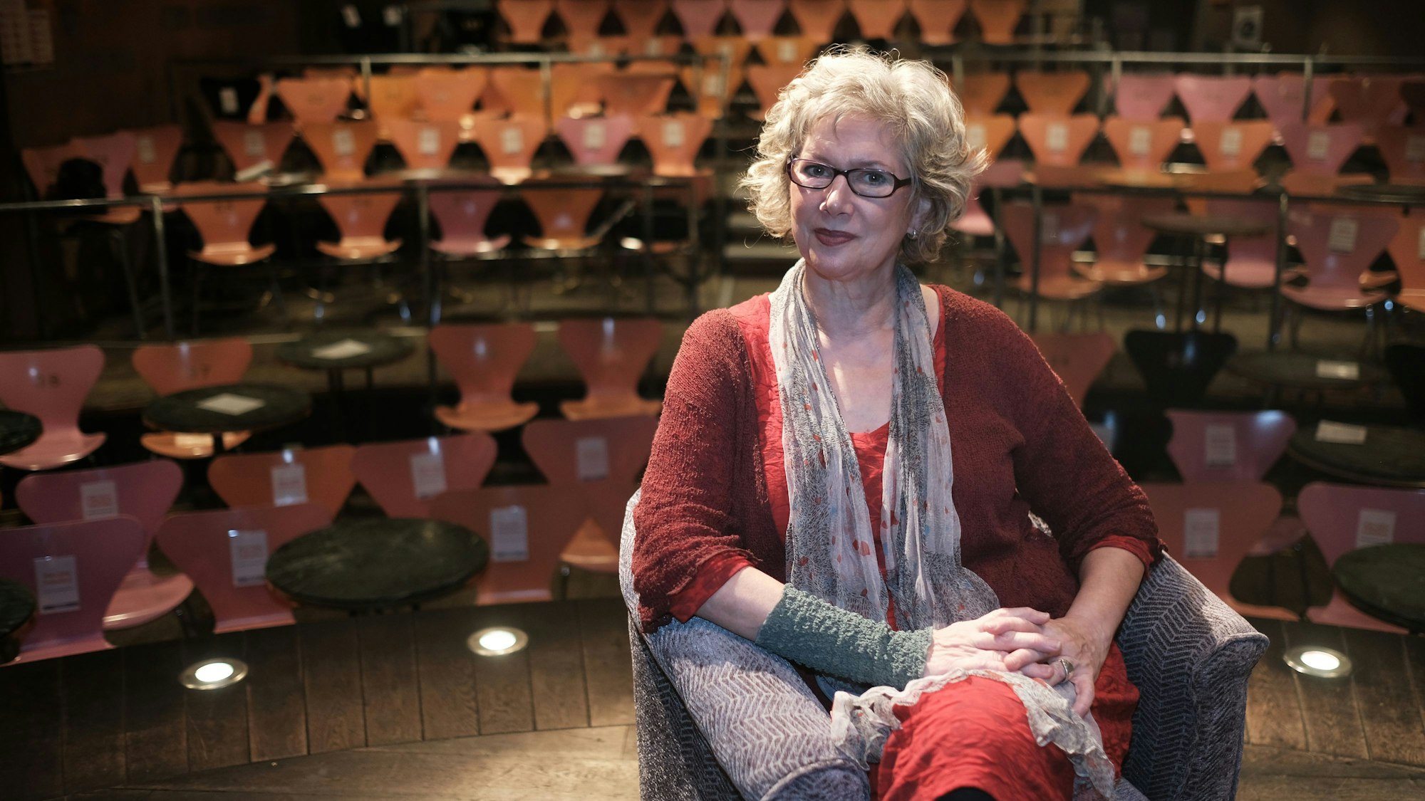 Eine Frau mittleren Alters sitzt auf der Bühne in einem kleinen Theater vor Stuhlreihen.