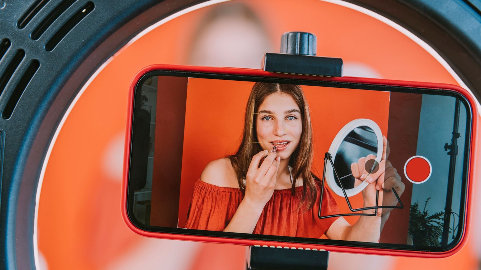 Auf einem Handy-Display sieht man das Bild einer Influencerin, die einen Spiegel in der Hand hält und sich die Lippen schminkt.
