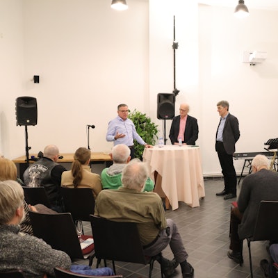 Drei Männer stehen bei einem Vortrag vor Publikum an einem Stehtisch.