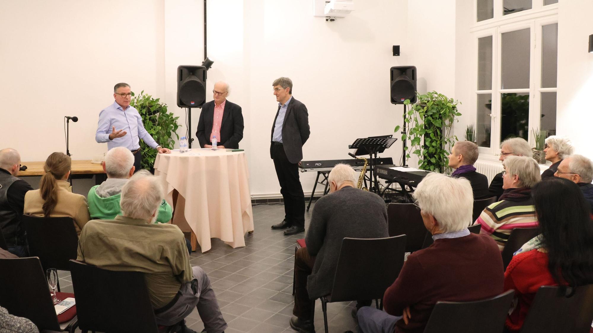Drei Männer stehen bei einem Vortrag vor Publikum an einem Stehtisch.
