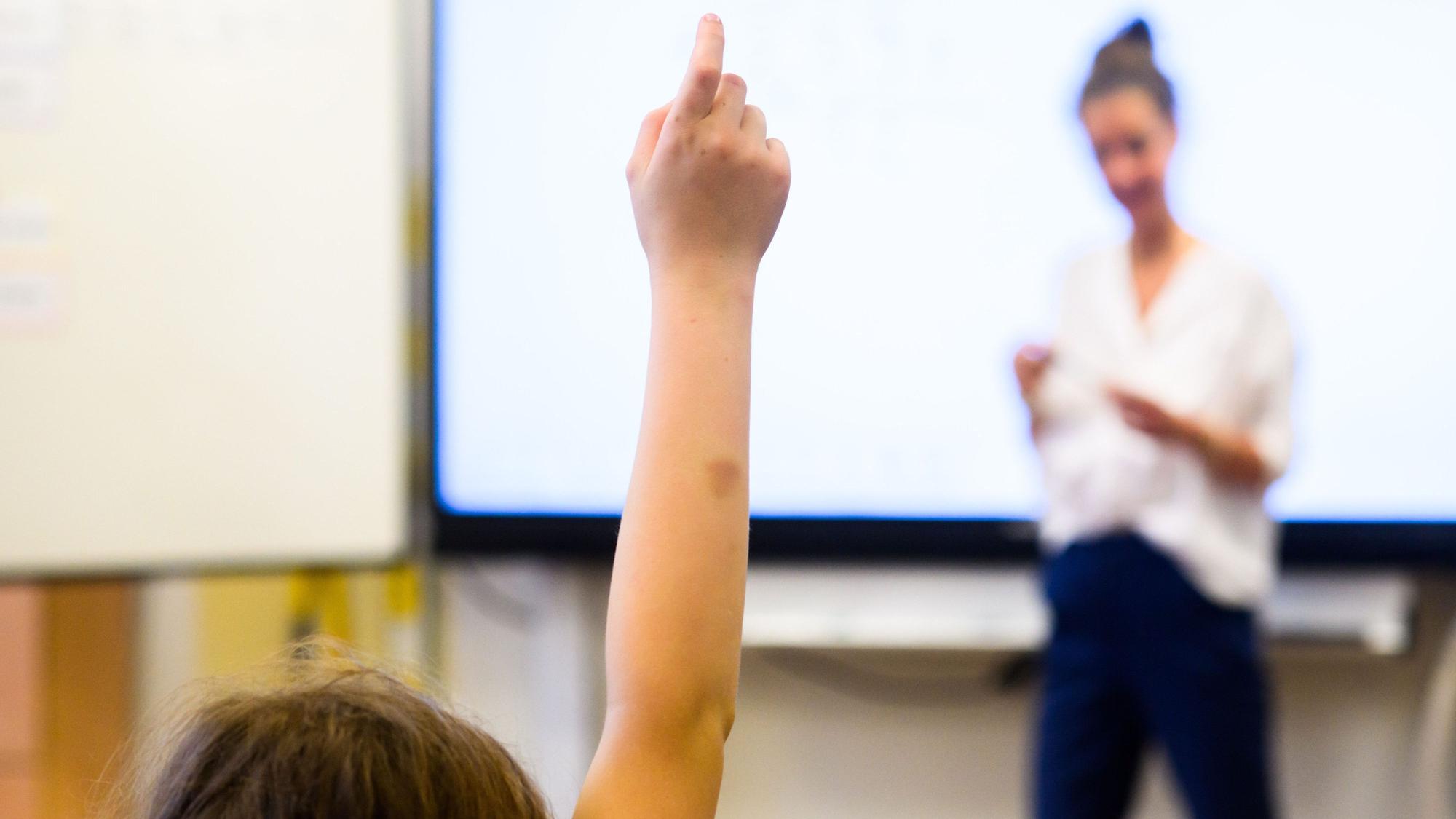 Ein Schüler meldet sich per Handzeichen, während eine Lehrerin vor einer digitalen Schultafel im Klassenraum steht.