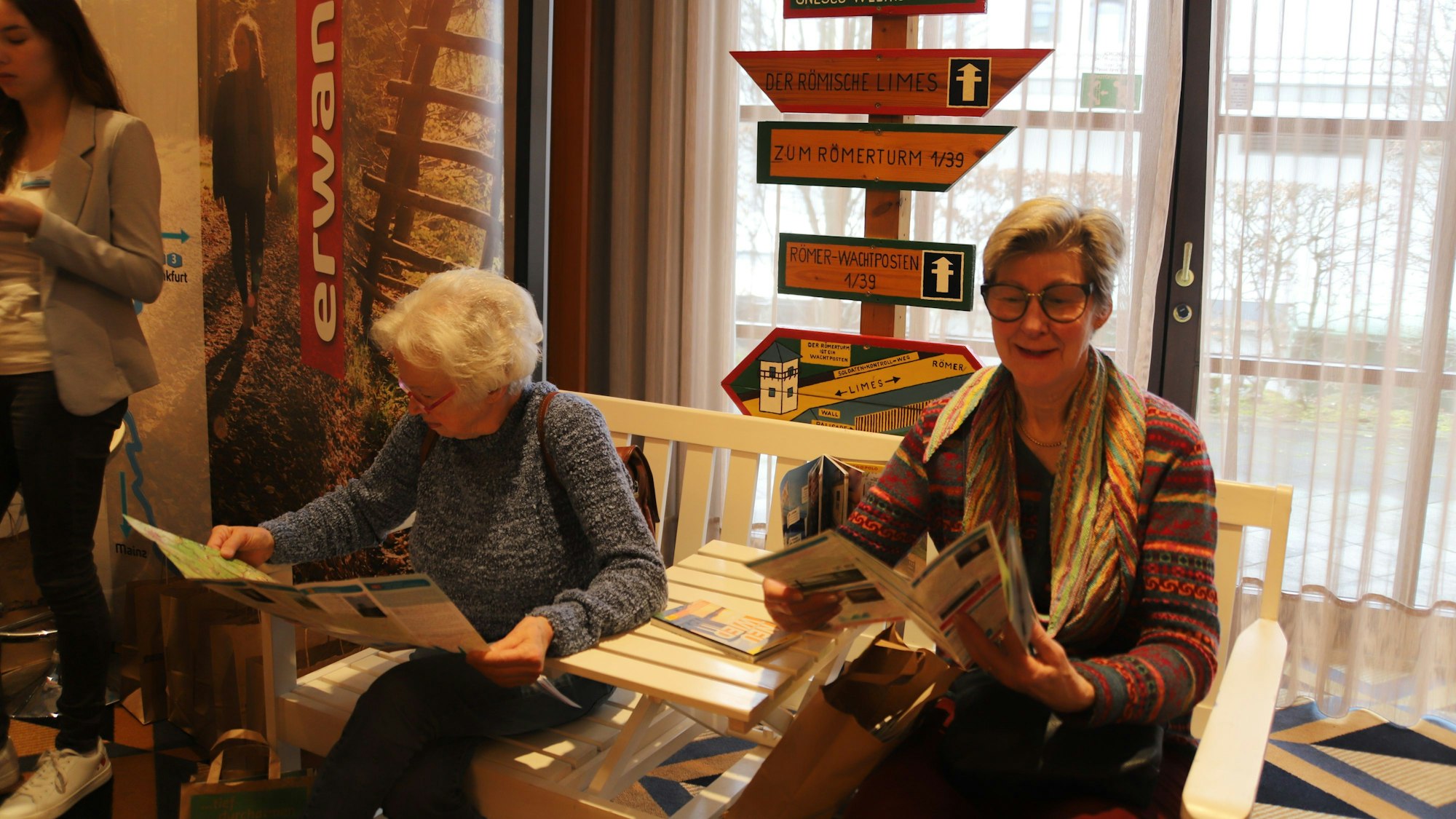 Zwei ältere Damen sitzen auf einer weiß lackierten Bank. Sie studieren Informationsbroschüren; hinter den beiden steht ein Wegweiser.