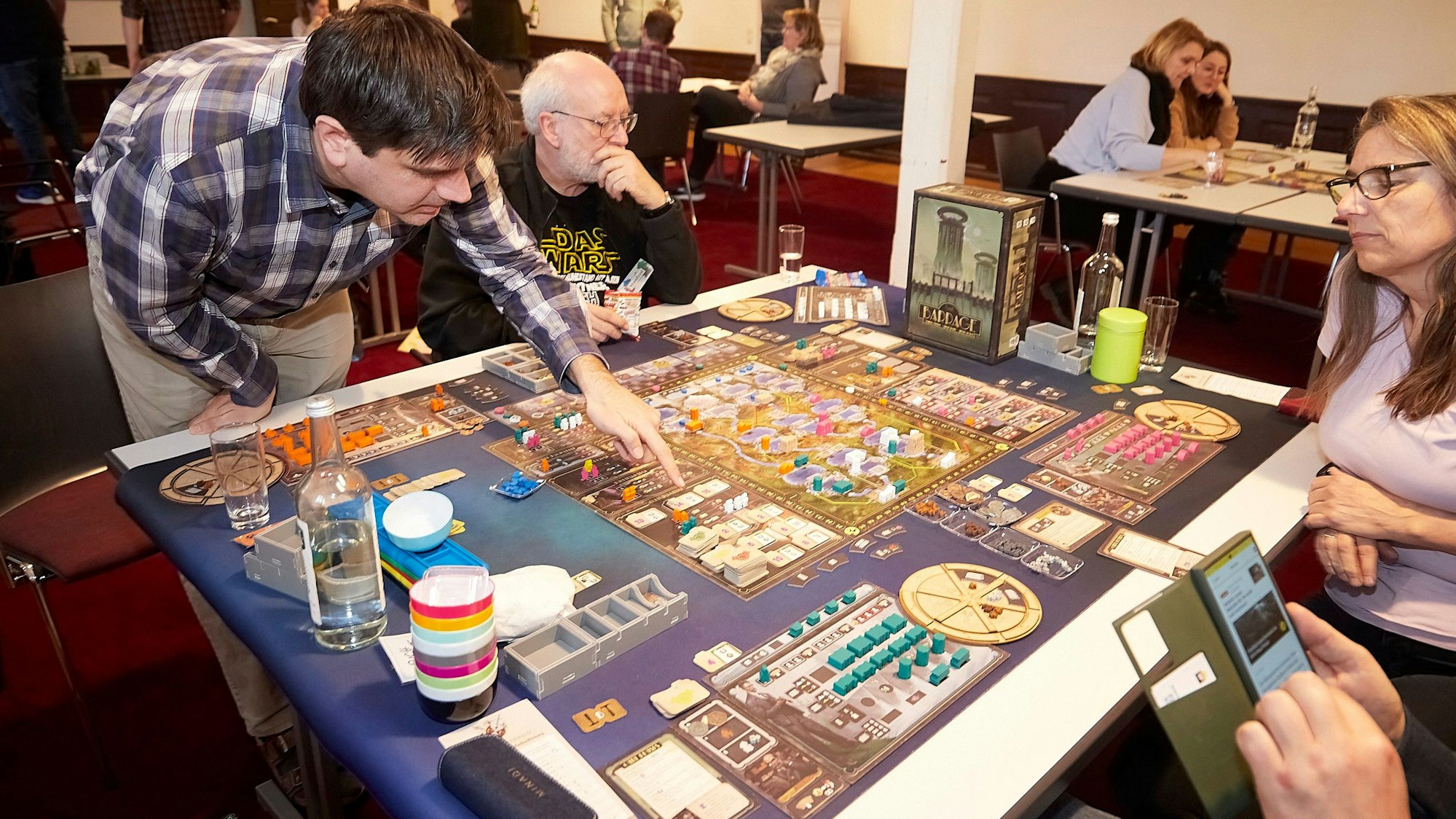 Vier Brettspieler sitzen um ein komplex aussehendes Spiel an einem Tisch. Einer von ihnen erklärt.