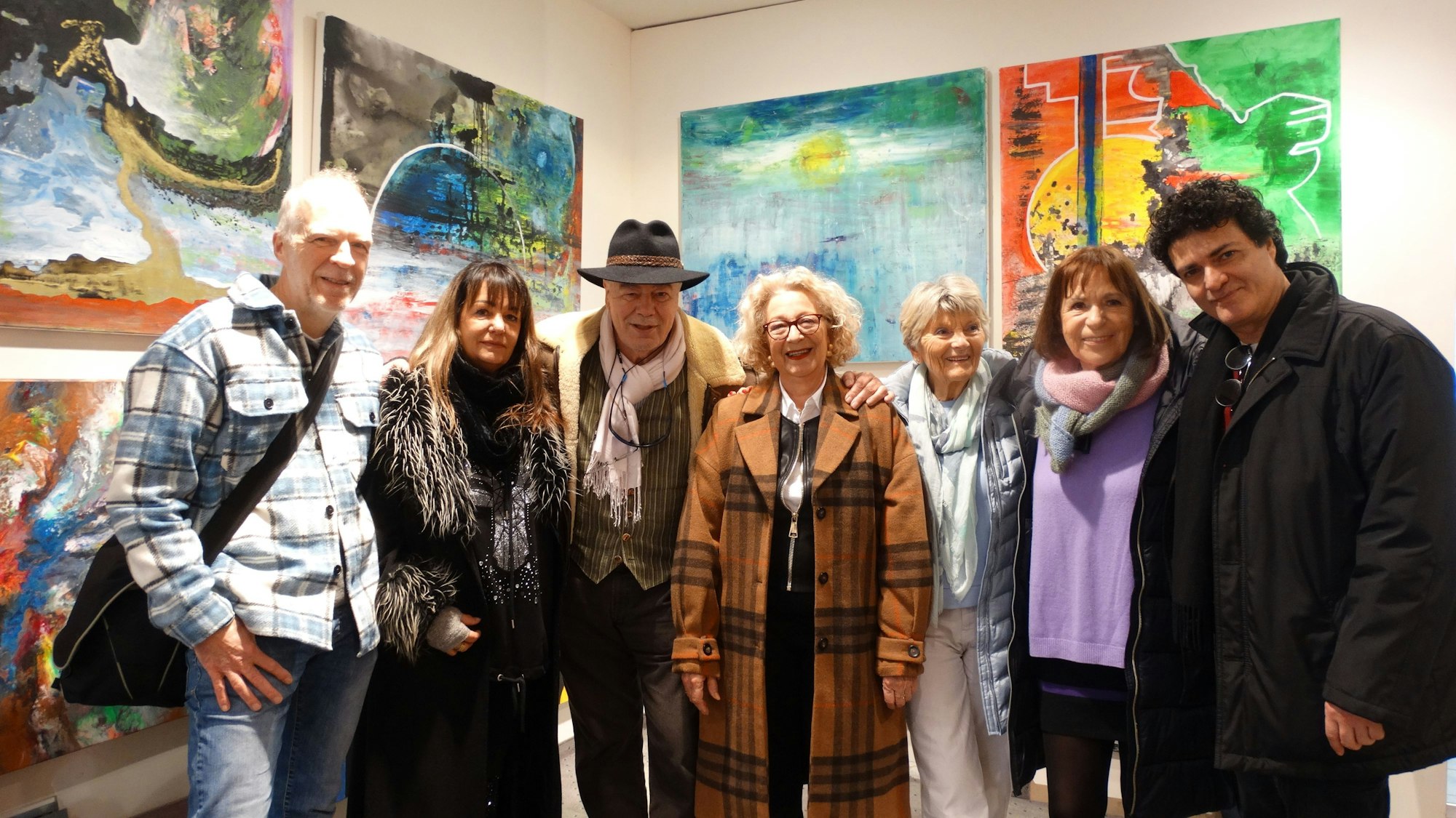 Männer und Frauen stehen vor Wänden, an denen bunte Gemälde hängen.