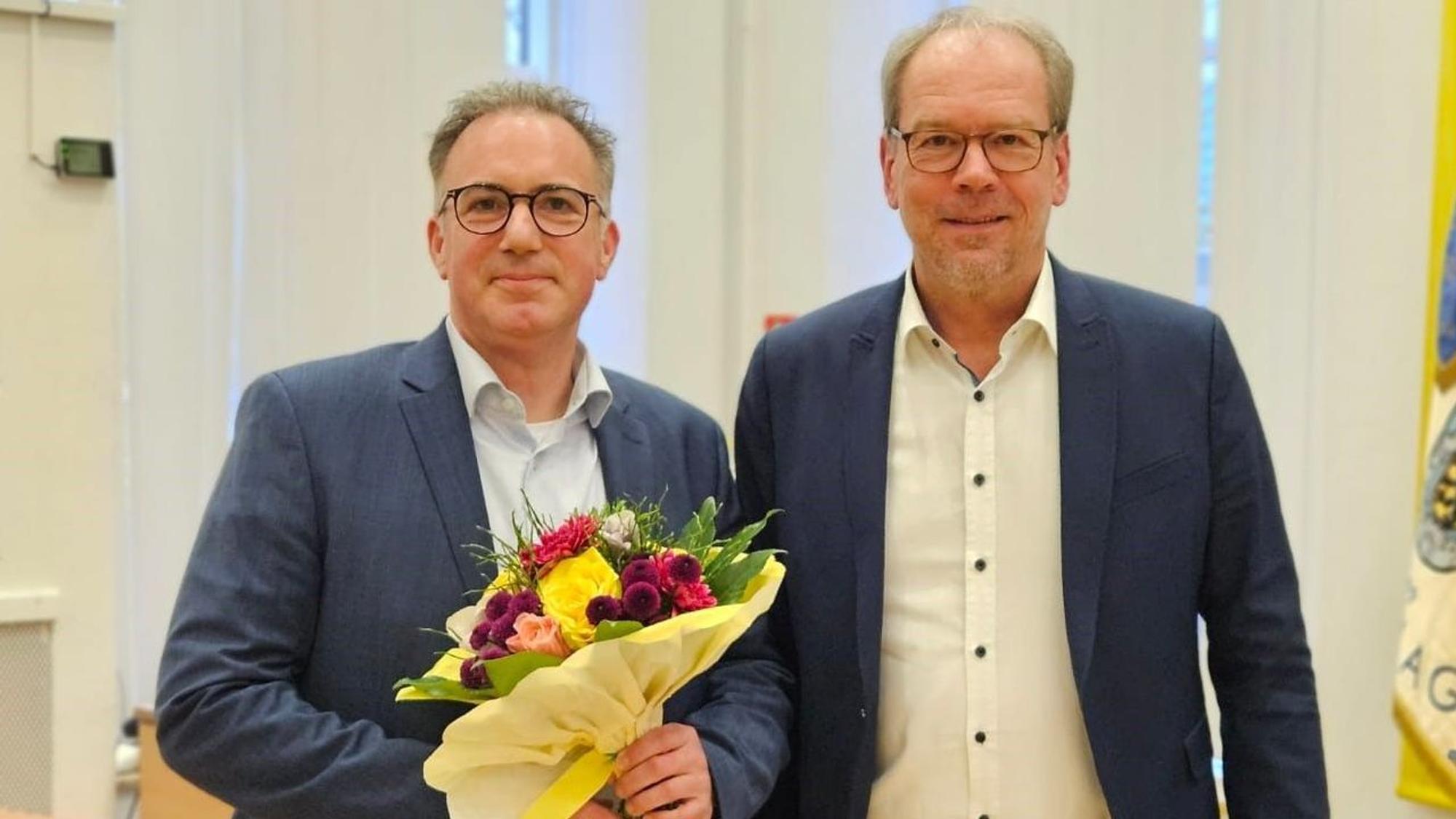 Zwei Männer lächeln, einer hält einen Blumenstrauß.
