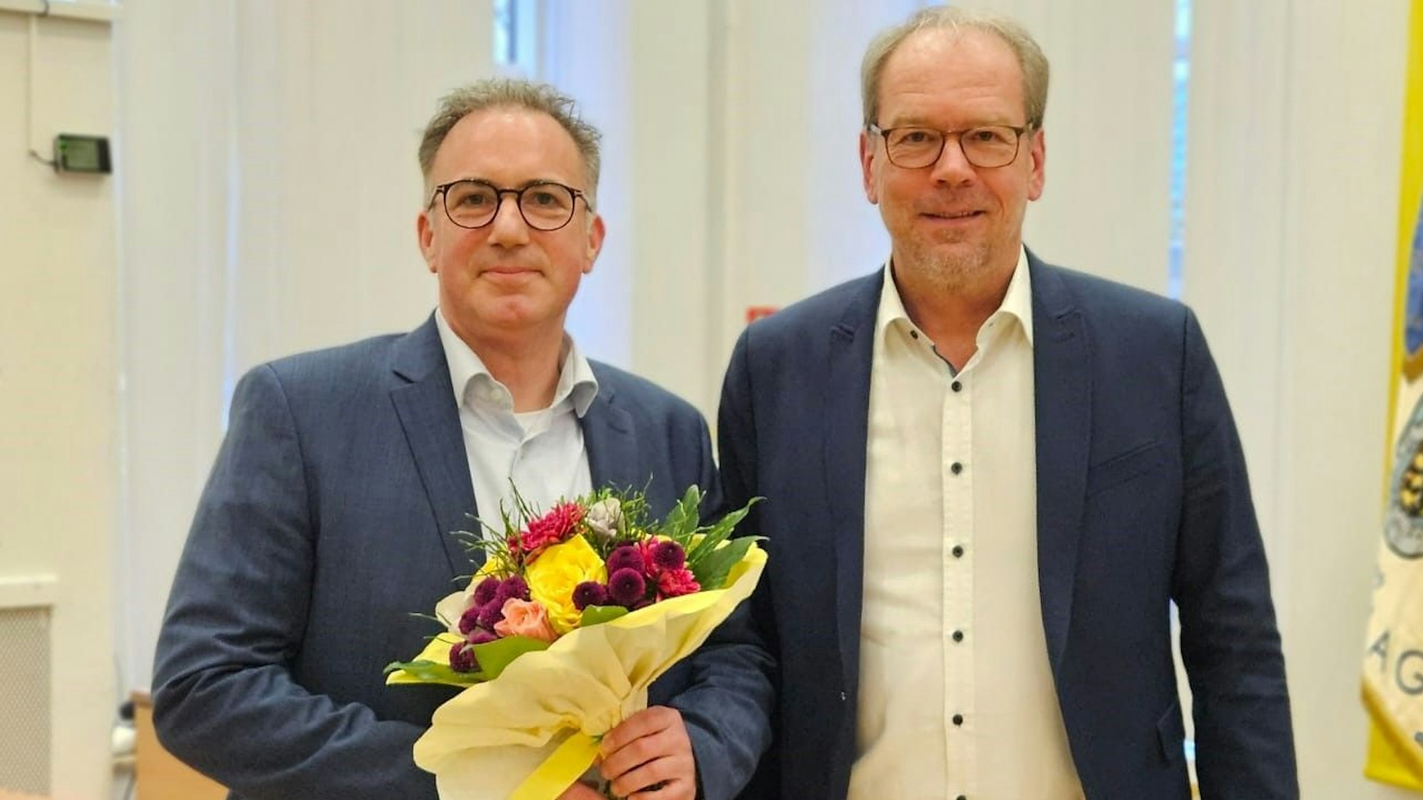 Zwei Männer lächeln, einer hält einen Blumenstrauß.