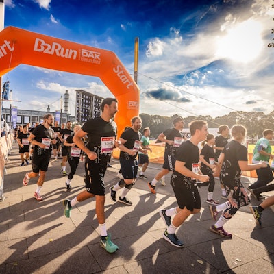 Zu sehen sind in schwarz gekleidete Menschen, die bei Sonnenschein joggen. Im Hintergrund befindet sich das Rhein-Energie-Stadion.