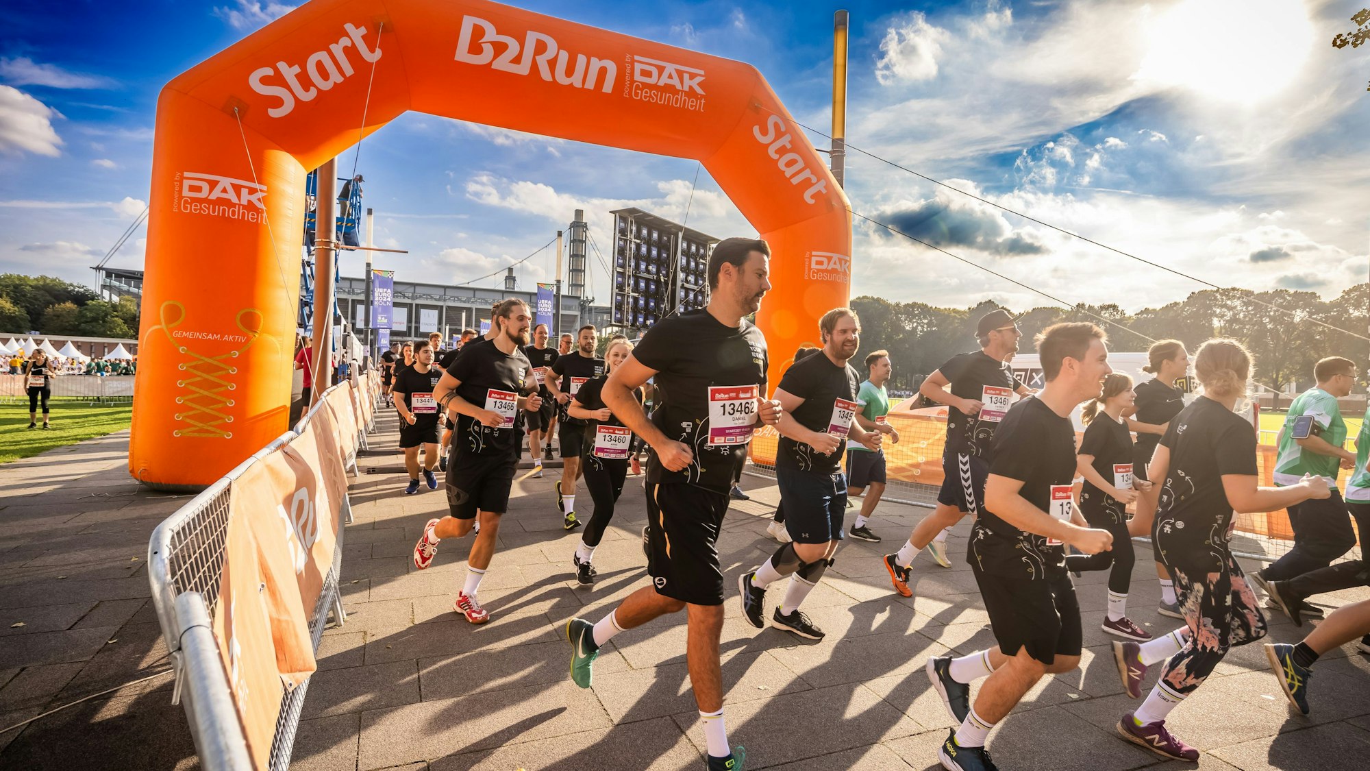 Zu sehen sind in schwarz gekleidete Menschen, die bei Sonnenschein joggen. Im Hintergrund befindet sich das Rhein-Energie-Stadion.