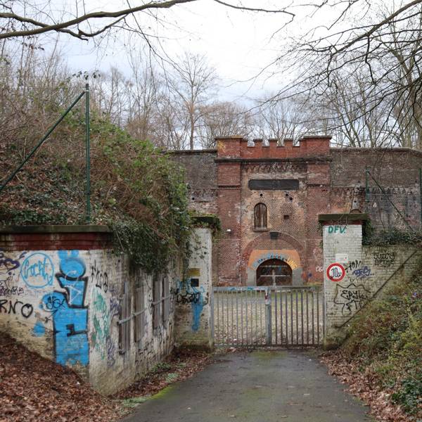 Blick auf eine Festungsanlage, die hinter einem verschlossenen Tor umgeben von Bäumen und Büschen liegt