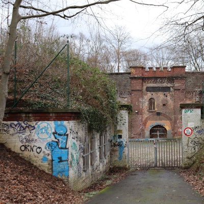 Blick auf eine Festungsanlage, die hinter einem verschlossenen Tor umgeben von Bäumen und Büschen liegt
