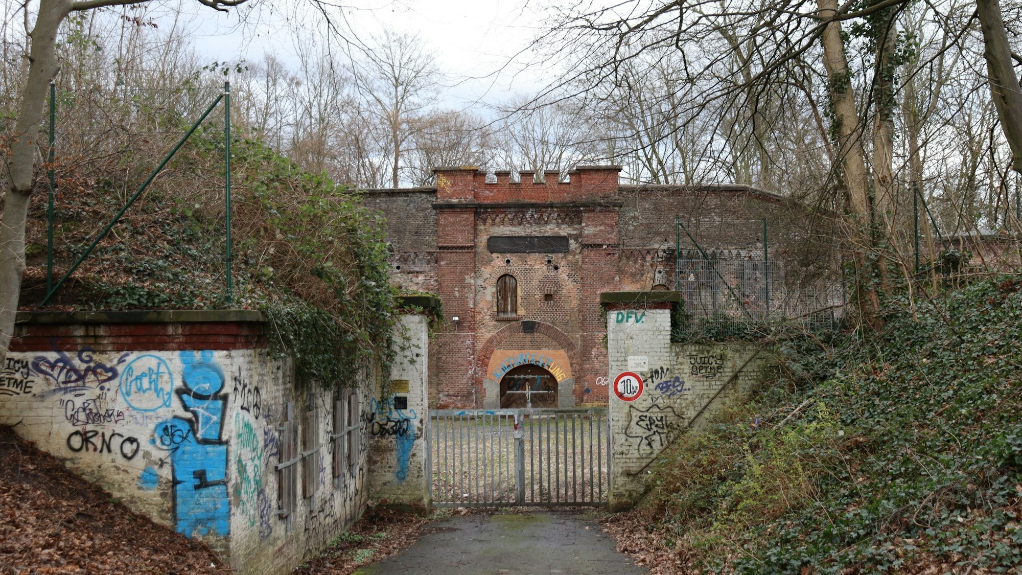 Blick auf eine Festungsanlage, die hinter einem verschlossenen Tor umgeben von Bäumen und Büschen liegt