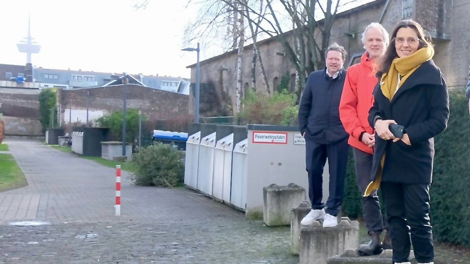Zwei Männer und eine Frau stehen auf niedrigen Betonpollern hintereinander. Im Hintergrund ist das Gebäude der Artilleriehalle zu sehen.