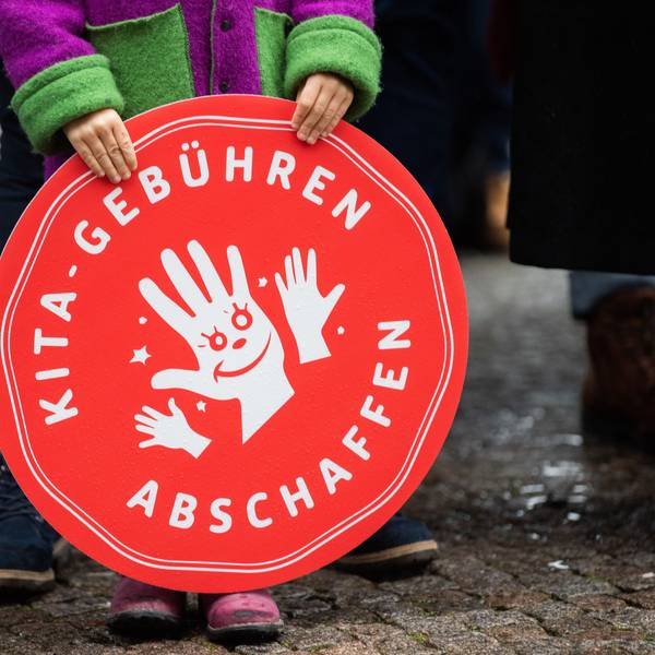 Ein Kind hält ein rotes, rundes Plakat mit der Aufschrift "Kita-Gebühren abschaffen" vor sich.