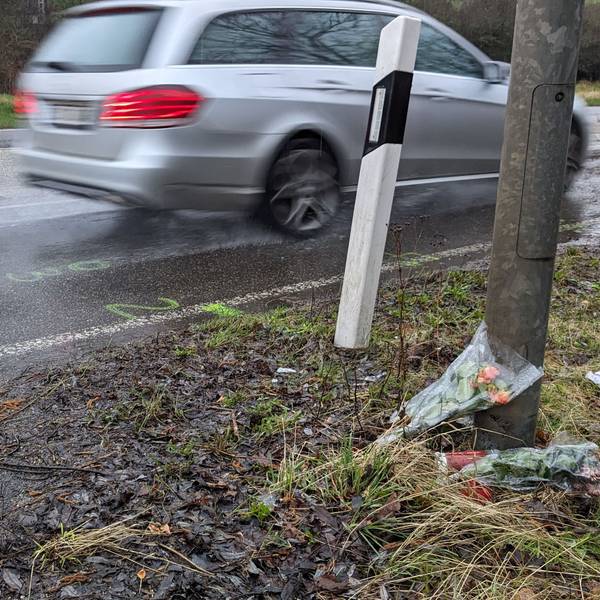 Am Straßenrand wurden an einem Baum Blumen und Kerzen abgelegt. Ein Auto fahrt vorbei.