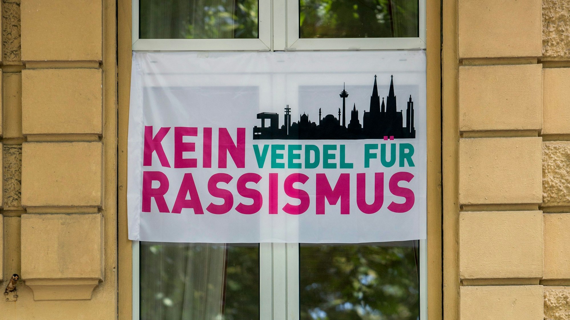 Fahnen der Initiative ‚Kein Veedel für Rassismus‘ hängt vor einem Fenster an einer Hauswand.