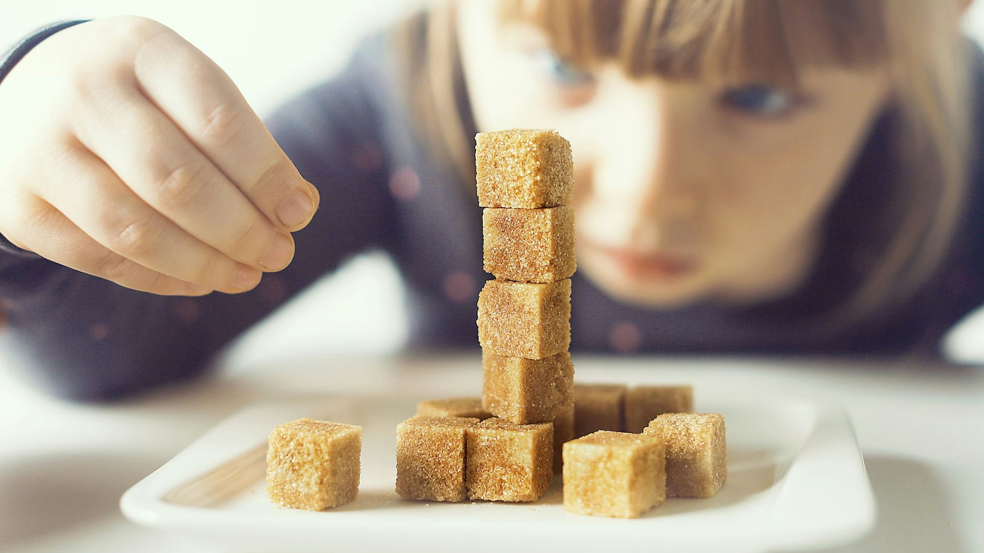 Ein kleines Mädchen sitzt an einem Tisch und stapelt Zuckerwürfel aufeinander.