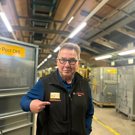 Ein älterer Mann mit Brille steht in einer Paketlagerhalle der Deutschen Post.