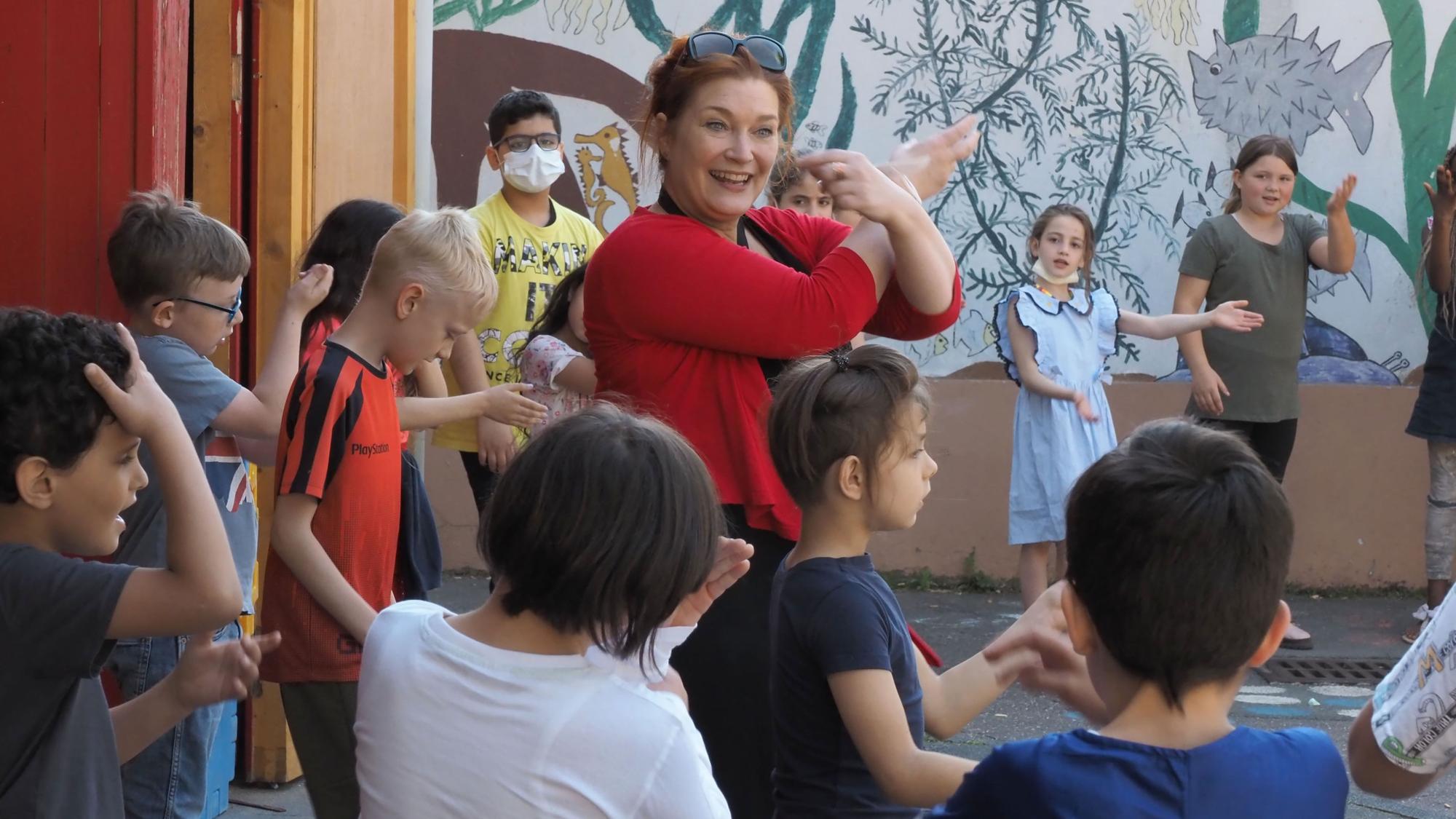Die Singleiterin Tanja Heesen vom Verein „Singpause Köln e.V.“ steht mit Grundschülerinnen und Grundschülern der Sternsingerschule in Longerich auf dem Pausenhof und gestikuliert wild.