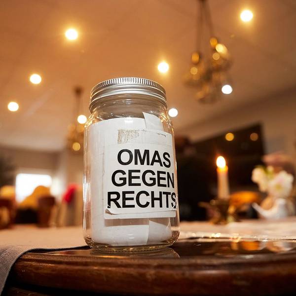 Ein Glas mit der Aufschrift Omas gegen Rechts steht auf einem Tisch. Im Hintergrund sind eine brennende Kerze und Menschen an weiteren Tischen zu erahnen.