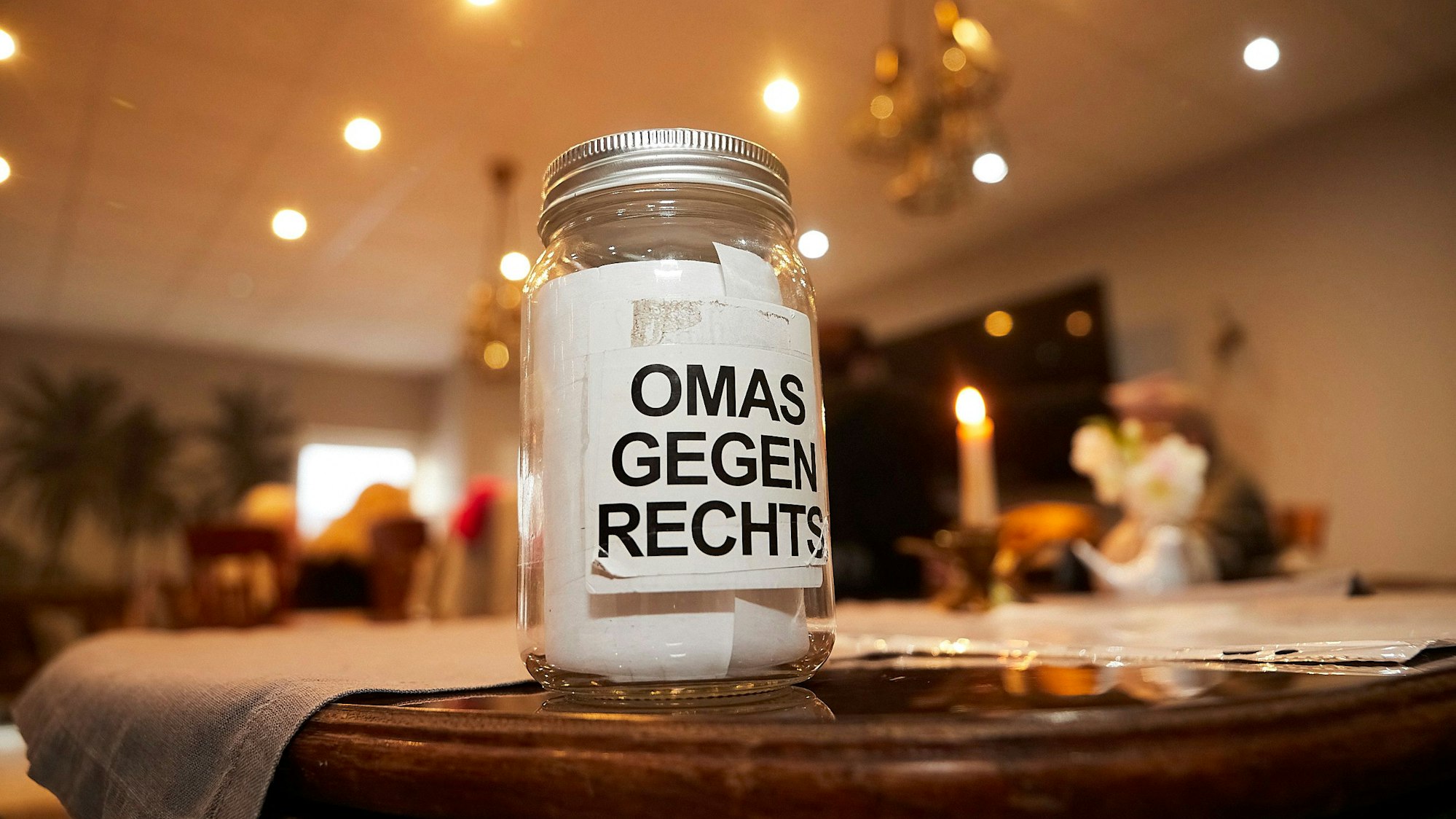 Ein Glas mit der Aufschrift Omas gegen Rechts steht auf einem Tisch. Im Hintergrund sind eine brennende Kerze und Menschen an weiteren Tischen zu erahnen.