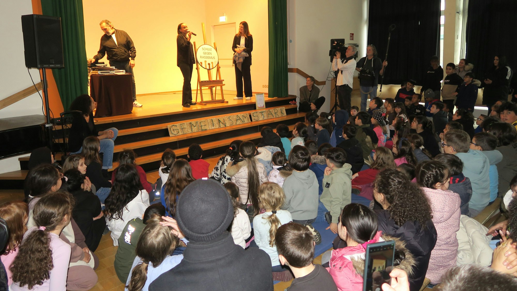 Auf einer Bühne stehen ein Mann und zwei Frauen. In der Schulaula sitzen viele Kinder und schauen zu.