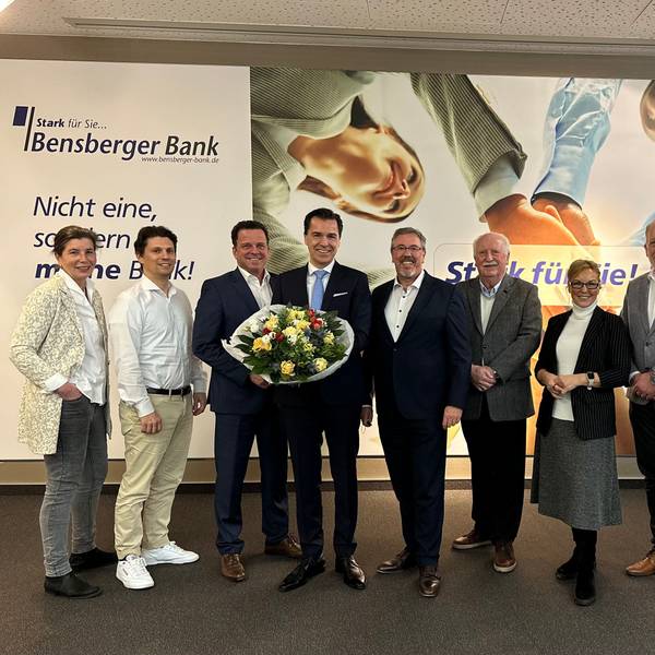 Vorstand und Aufsichtsrat der Bensberger Bank stehen vor einer Reklame der Genossenschaftsbank.